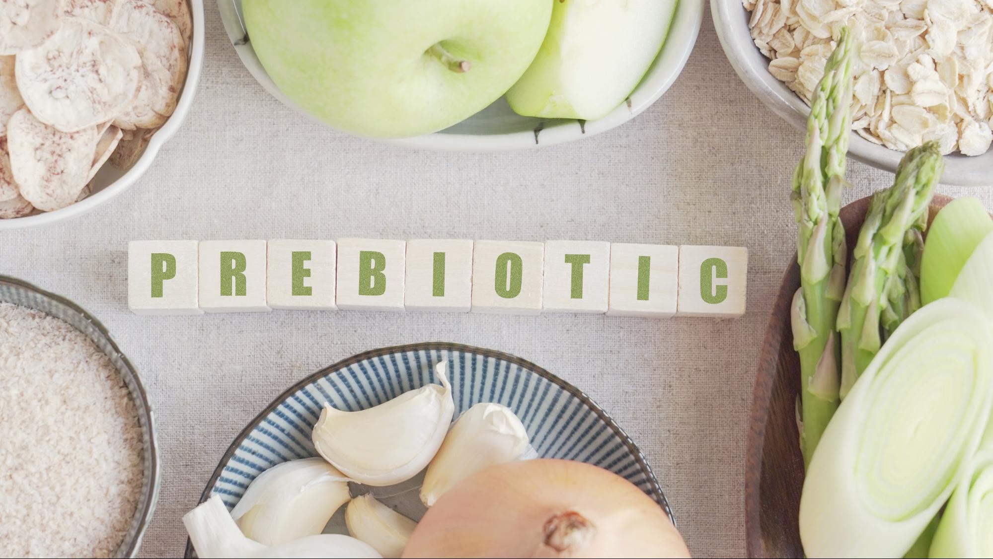 The word prebiotic spelled out in wooden blocks surrounded by prebiotic foods including garlic, onions, apples, and leeks.